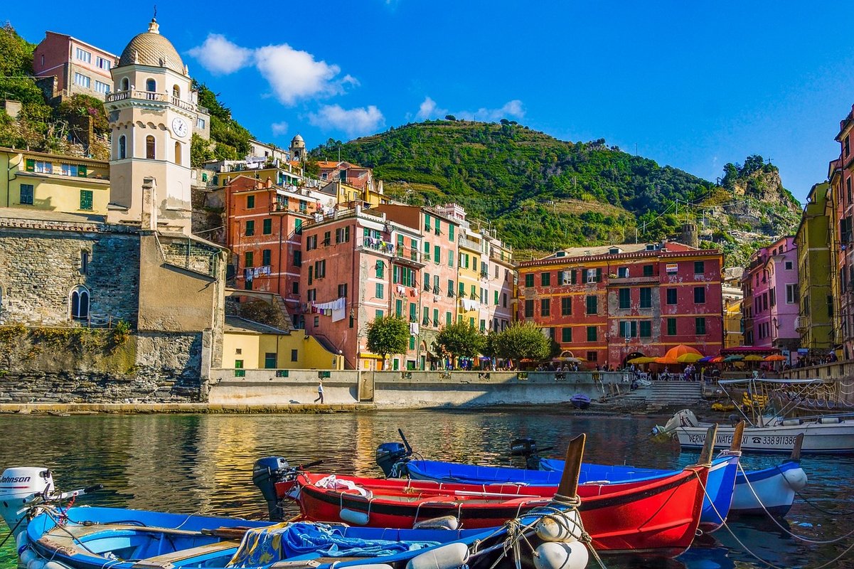 Ville italienne avec au premier plan un plan d'eau et des barques colorées, et en fond une ville entourée de montagnes.