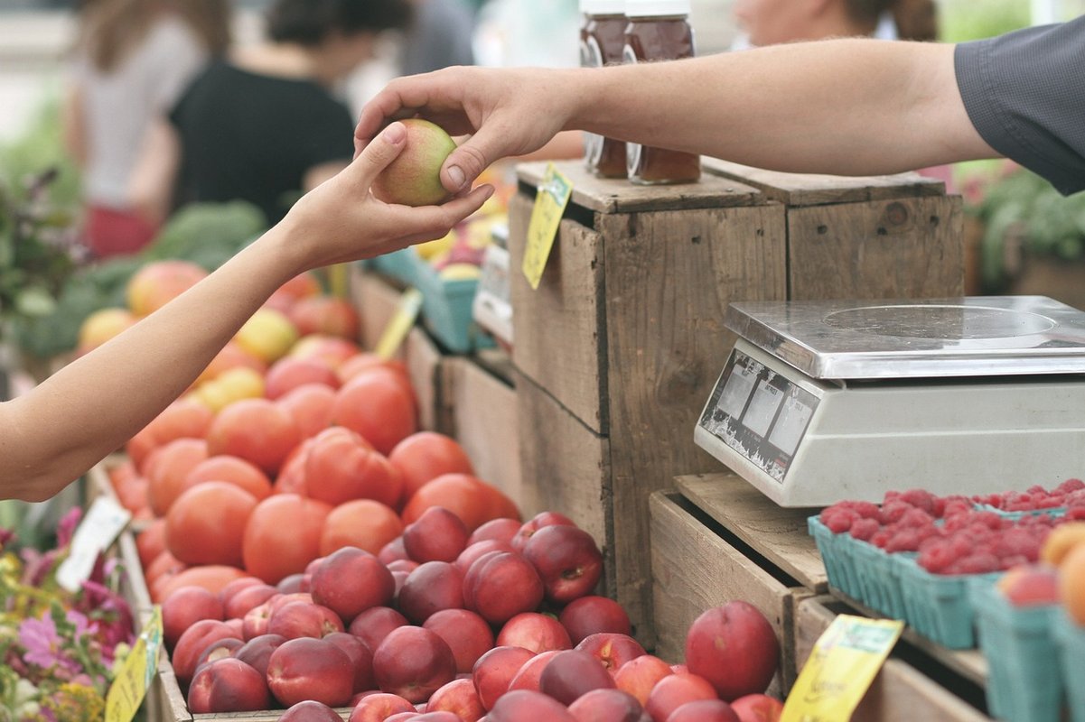 On y voit deux mains échanger un fruit avec en fond une étale de marché